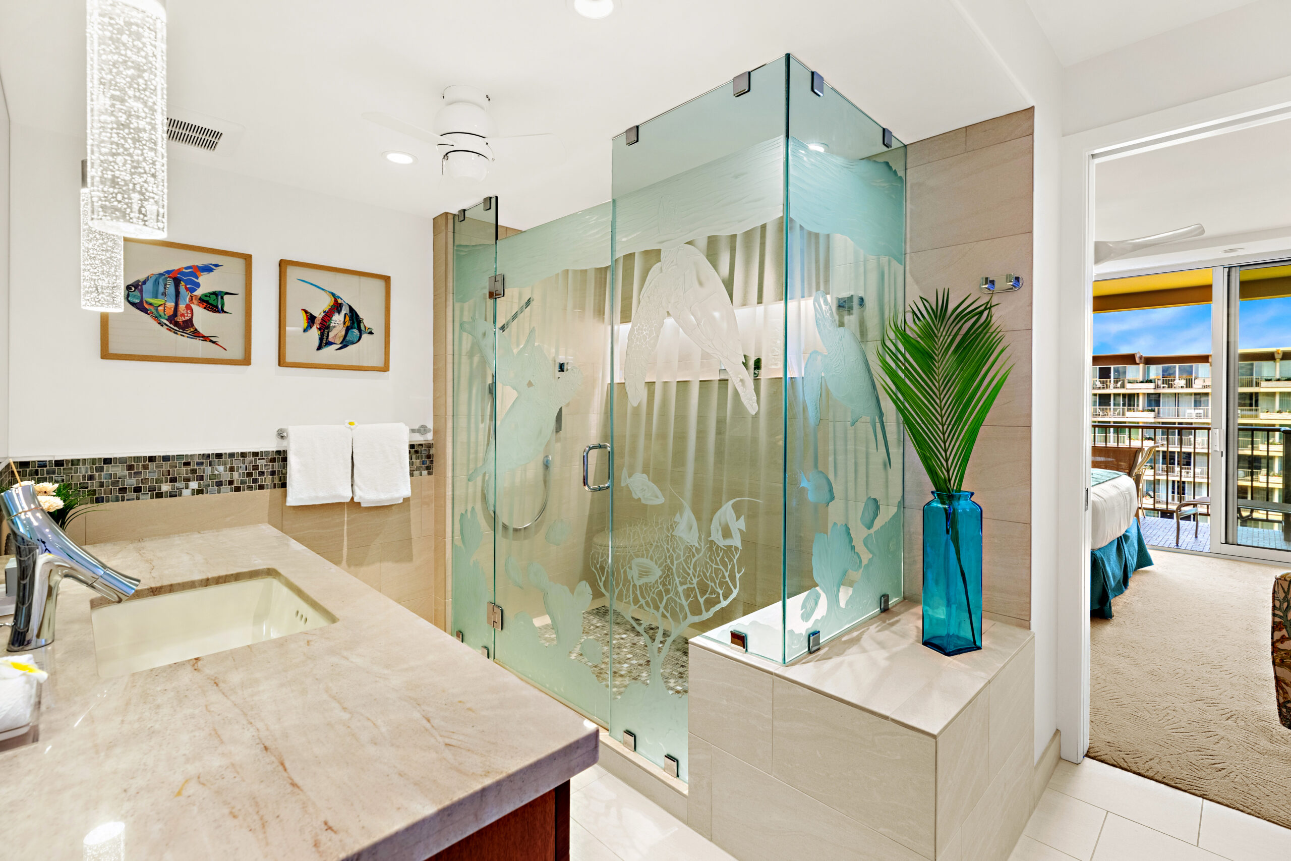 Modern bathroom with glass shower featuring ocean designs, double sink, two fish paintings, and a blue vase with a palm leaf. Bedroom and balcony visible.