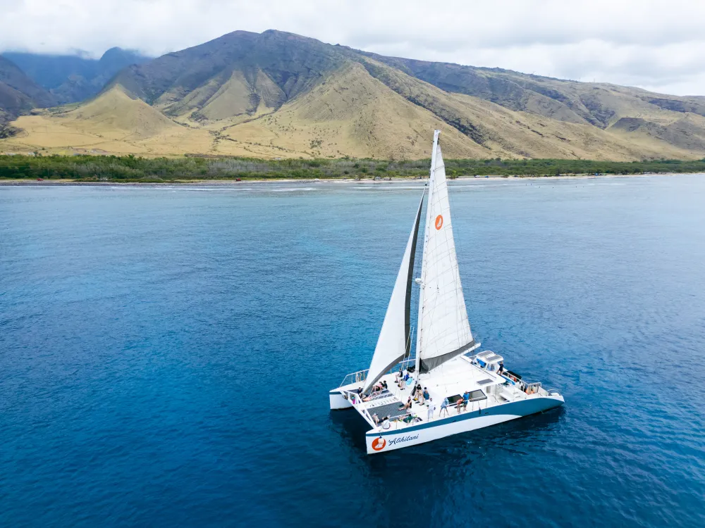 A white catamaran sails on calm blue water near Kaanapali’s green mountains—perfect for group rentals under a cloudy sky.