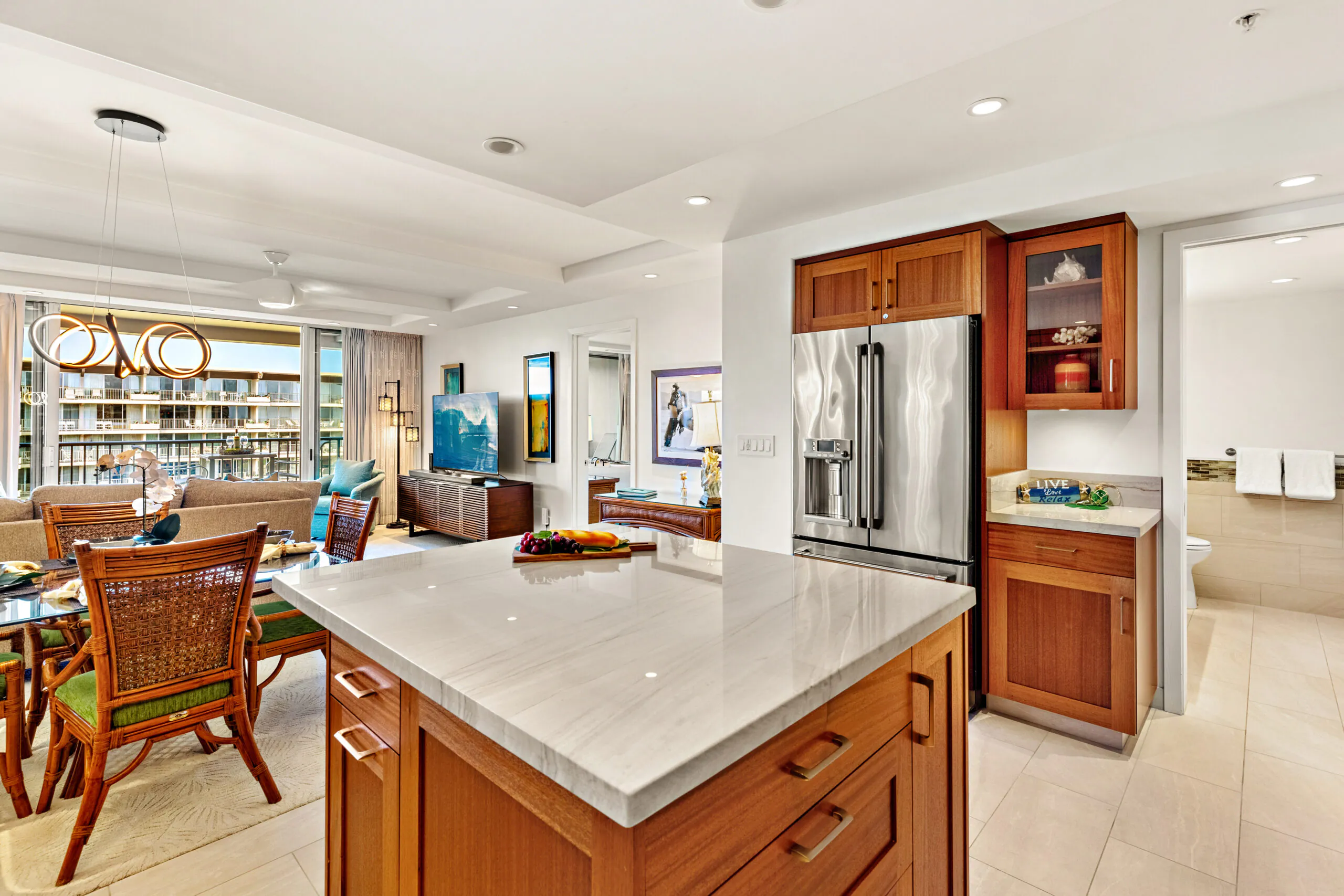 Modern kitchen with wood cabinets, large island, and stainless steel fridge, open to a bright living room and balcony with outdoor view.