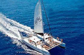 A white catamaran with people on board sails across clear blue ocean water on a sunny day, leaving a wake behind.