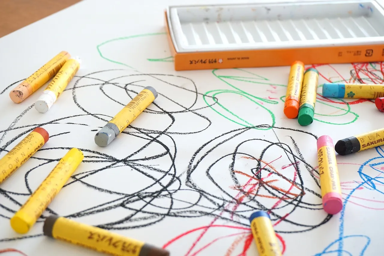Crayons and a box lie on paper covered with colorful, scribbled drawings in black, green, and red.