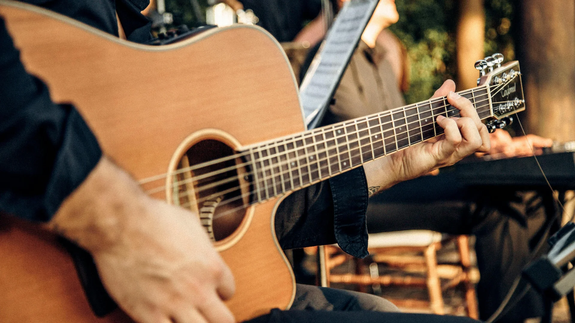 Close-up of a person playing an acoustic guitar outdoors, with sunlight and blurred musicians in the background.
