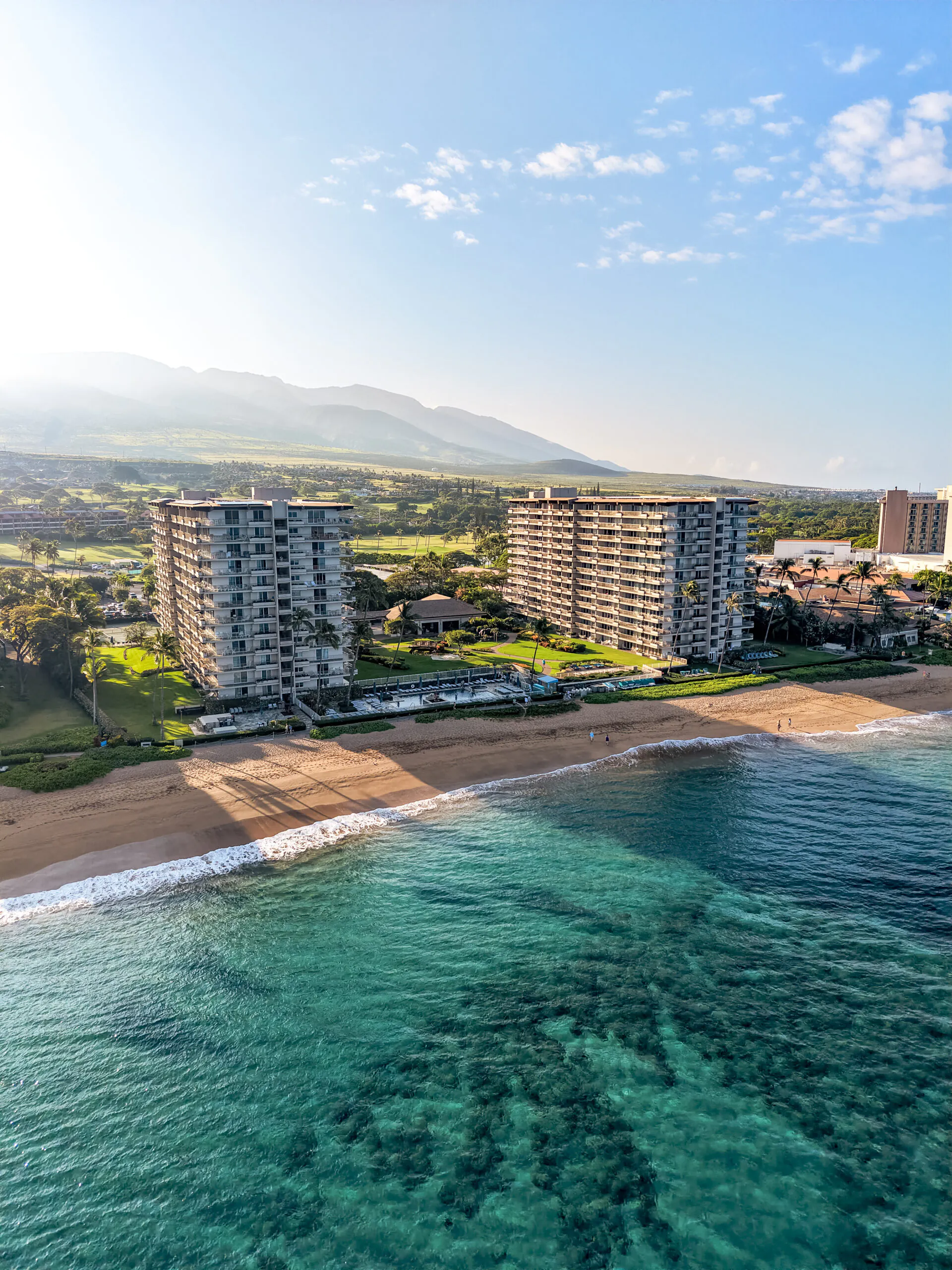 Aerial view of beachfront condos with lush greenery, sandy shore, and clear turquoise water under a sunny sky with mountains in the background.