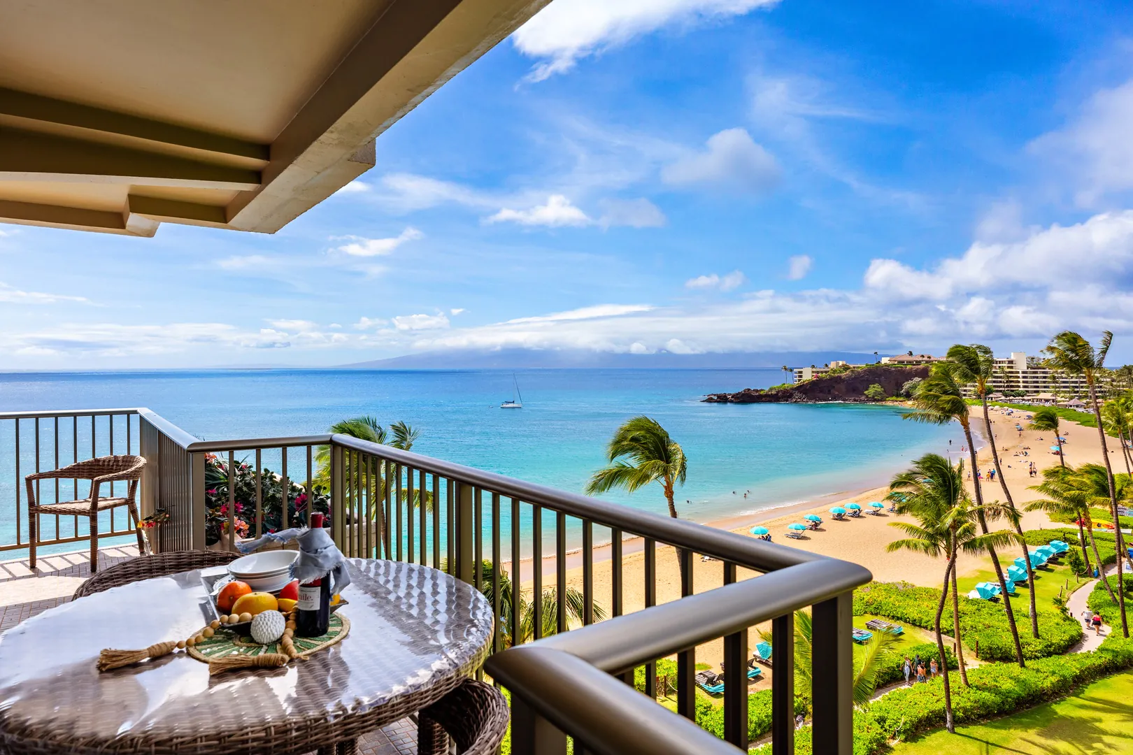 A balcony at Kaanapali HI beachfront resorts overlooks a sandy beach with palm trees, blue umbrellas, and ocean views. Drinks and fruit await on the table.